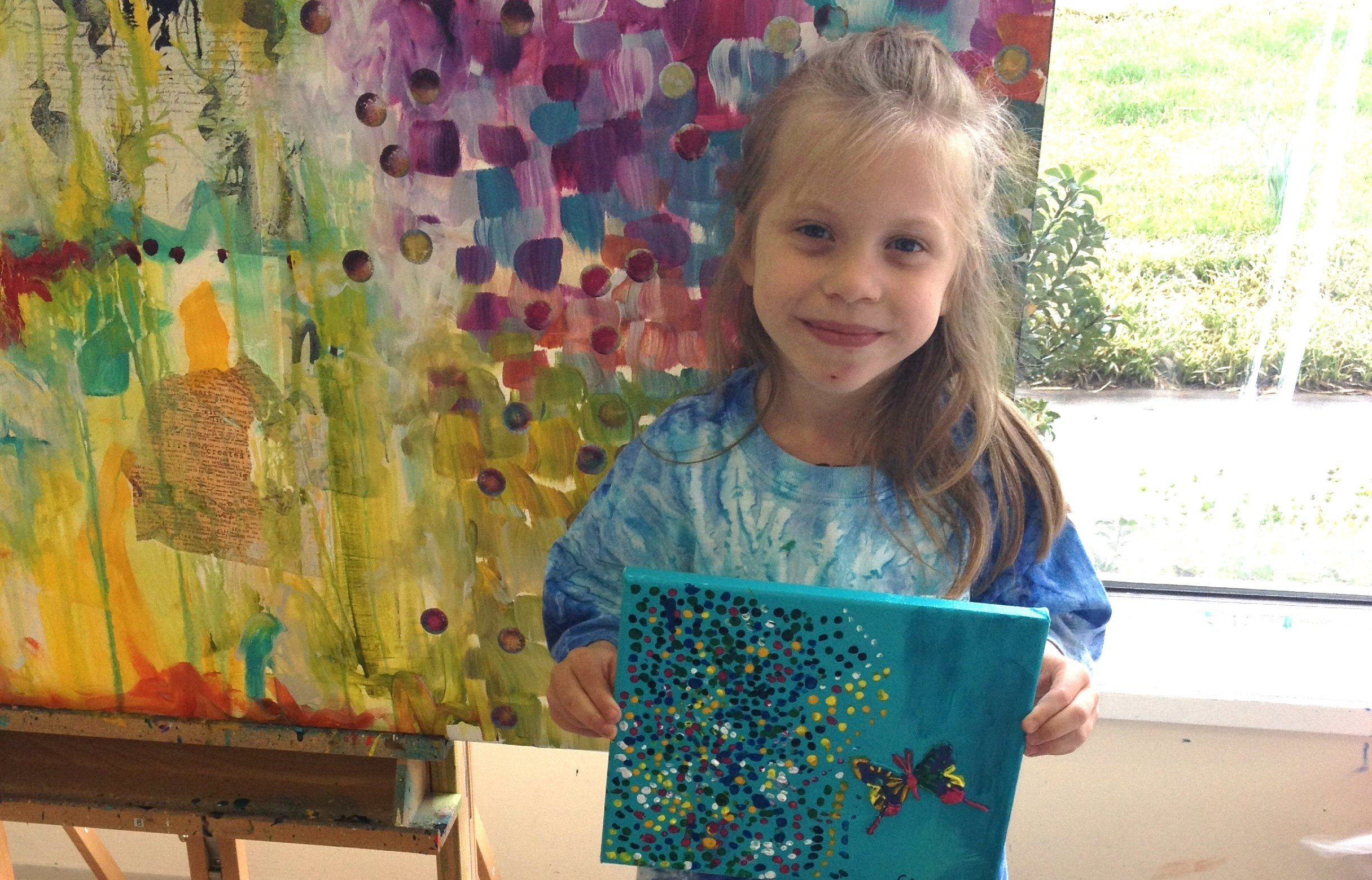 A little girl holds a blue painting with a butterfly and dots while standing in front of a colorful painting.